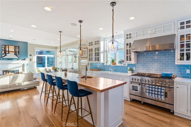 kitchen featuring exhaust hood, decorative light fixtures, double oven range, an island with sink, and white cabinets