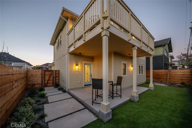 back house at dusk with a balcony, a lawn, and a patio area