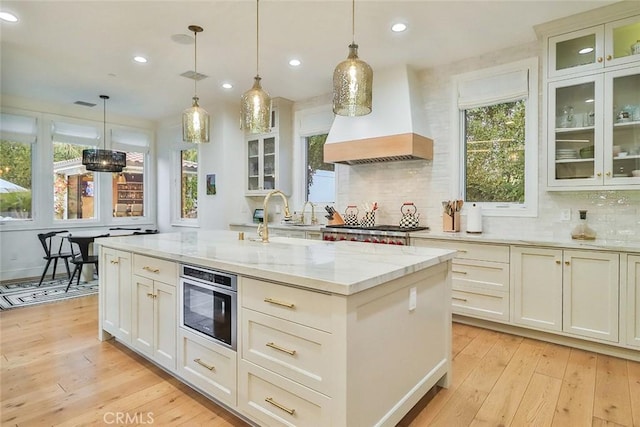 kitchen with custom exhaust hood, light hardwood / wood-style floors, an island with sink, hanging light fixtures, and light stone countertops