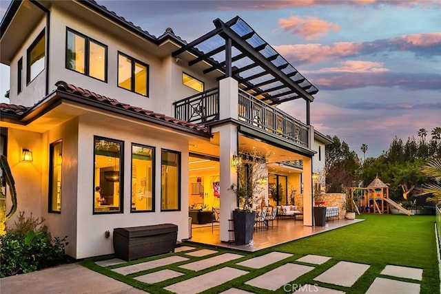 back house at dusk with a playground, a balcony, a lawn, and a patio