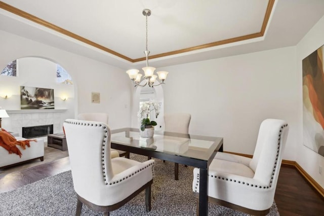 dining area with dark wood-type flooring, a notable chandelier, a tray ceiling, ornamental molding, and a tiled fireplace