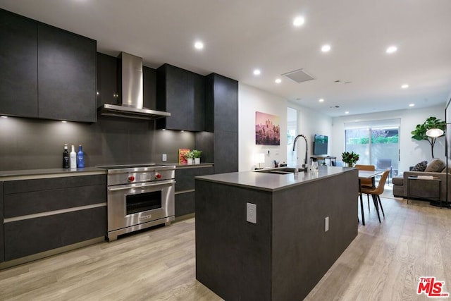 kitchen with wall chimney range hood, an island with sink, light hardwood / wood-style floors, sink, and premium stove