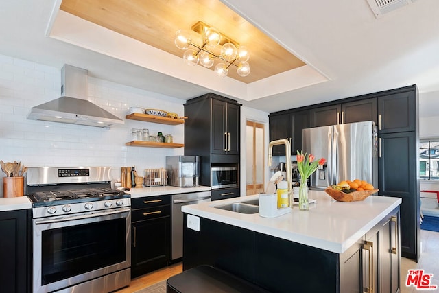 kitchen featuring sink, a tray ceiling, appliances with stainless steel finishes, an island with sink, and wall chimney exhaust hood