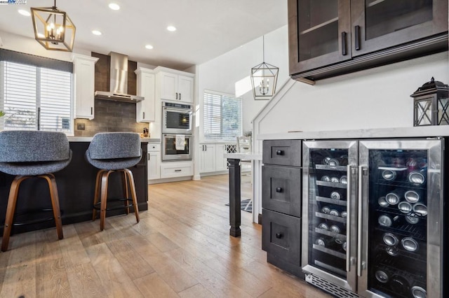 kitchen featuring decorative backsplash, white cabinetry, hanging light fixtures, stainless steel double oven, and wall chimney exhaust hood