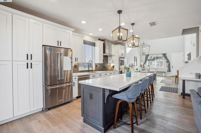 kitchen featuring a center island, wall chimney exhaust hood, white cabinetry, stainless steel appliances, and hanging light fixtures