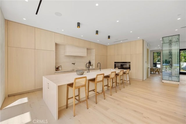 kitchen with a center island with sink, black oven, light brown cabinets, light wood-type flooring, and a breakfast bar