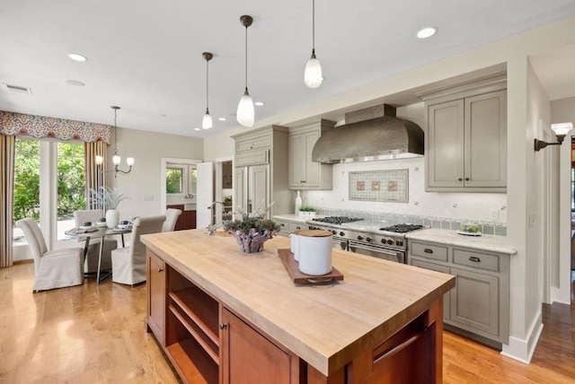 kitchen with wall chimney range hood, a center island with sink, double oven range, decorative backsplash, and hanging light fixtures