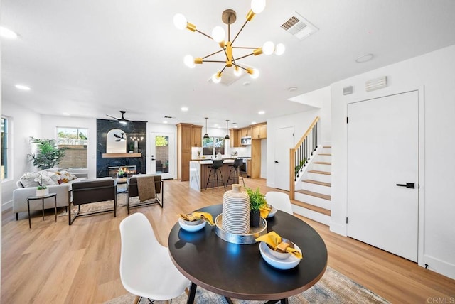 dining space featuring light wood-type flooring, ceiling fan with notable chandelier, and a fireplace