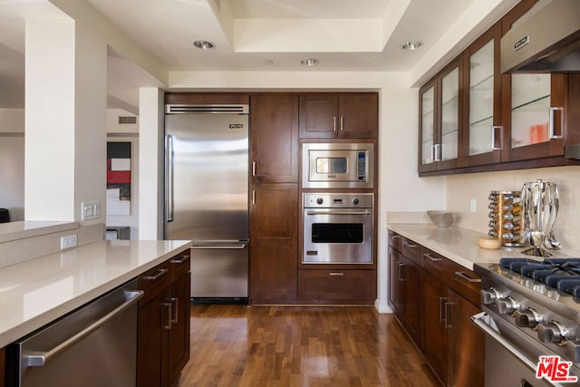 kitchen with exhaust hood, built in appliances, a tray ceiling, dark hardwood / wood-style flooring, and light stone counters