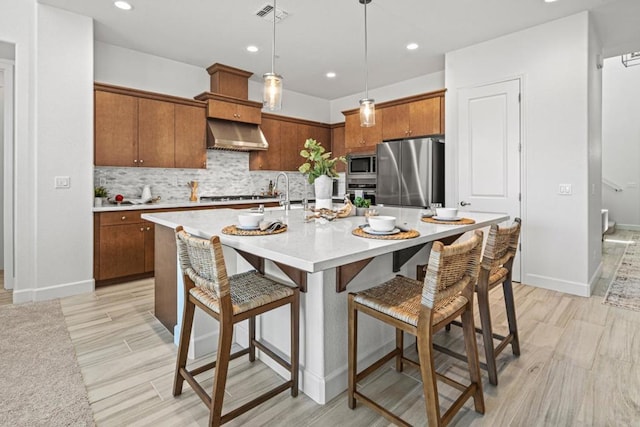 kitchen with appliances with stainless steel finishes, decorative light fixtures, wall chimney range hood, a kitchen breakfast bar, and a kitchen island with sink