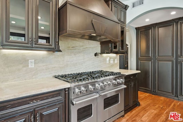 kitchen featuring backsplash, light hardwood / wood-style flooring, dark brown cabinetry, range with two ovens, and custom range hood