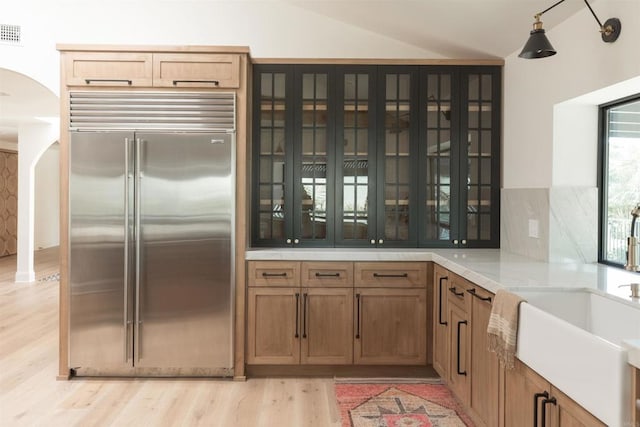 kitchen with glass insert cabinets, stainless steel built in fridge, arched walkways, lofted ceiling, and light wood-style floors