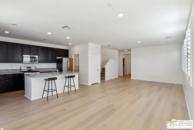 kitchen with an island with sink, appliances with stainless steel finishes, a kitchen breakfast bar, light stone countertops, and light hardwood / wood-style flooring