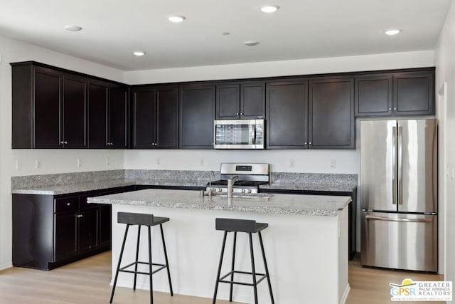 kitchen featuring dark brown cabinetry, light wood-type flooring, appliances with stainless steel finishes, an island with sink, and light stone counters