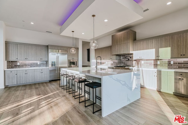 kitchen with decorative light fixtures, a large island with sink, light hardwood / wood-style floors, built in appliances, and light stone counters