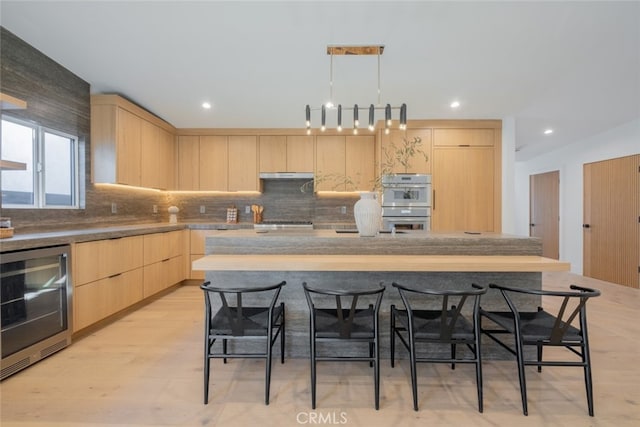 kitchen featuring tasteful backsplash, a kitchen island with sink, light brown cabinetry, beverage cooler, and double oven