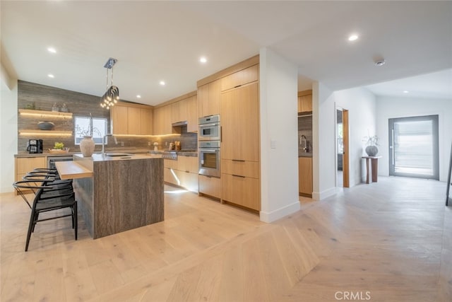 kitchen featuring pendant lighting, stainless steel double oven, light brown cabinetry, sink, and a kitchen island with sink