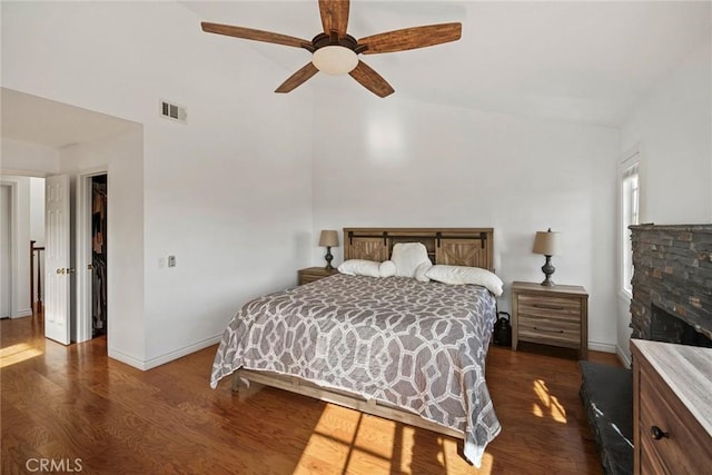 bedroom with a stone fireplace, ceiling fan, dark wood-type flooring, vaulted ceiling, and a walk in closet