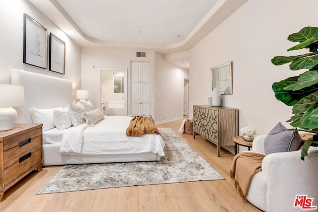 bedroom with ensuite bathroom, a raised ceiling, and light wood-type flooring