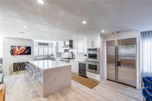 kitchen with white cabinetry, appliances with stainless steel finishes, a kitchen breakfast bar, beverage cooler, and wall chimney exhaust hood