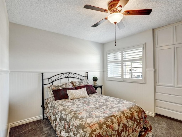 bedroom featuring a wainscoted wall, ceiling fan, dark colored carpet, and a textured ceiling