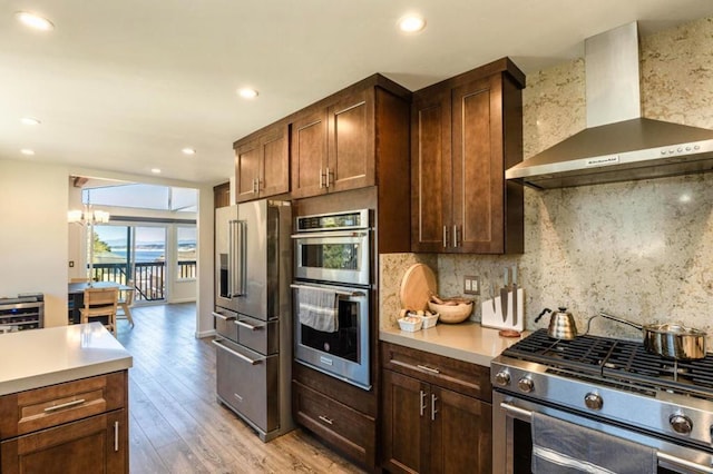 kitchen featuring appliances with stainless steel finishes, a chandelier, decorative backsplash, light hardwood / wood-style floors, and wall chimney exhaust hood