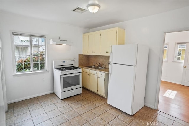 kitchen with range hood, cream cabinetry, a sink, white appliances, and plenty of natural light