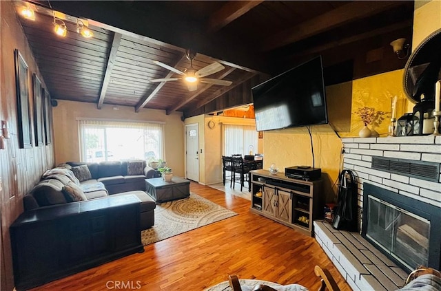 living room featuring vaulted ceiling with beams, wooden ceiling, wood finished floors, a ceiling fan, and a glass covered fireplace