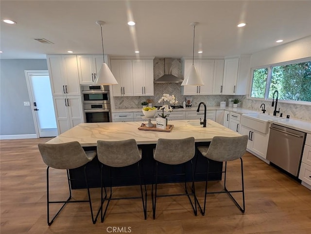 kitchen with stainless steel appliances, pendant lighting, a center island with sink, and wall chimney range hood