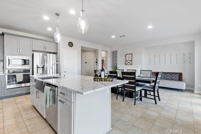 kitchen featuring stainless steel appliances, gray cabinetry, a kitchen island with sink, hanging light fixtures, and light stone countertops