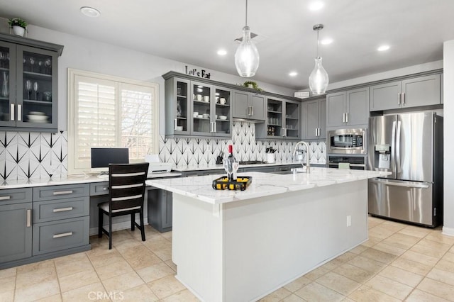 kitchen with stainless steel appliances, decorative light fixtures, a kitchen island with sink, built in desk, and light stone counters