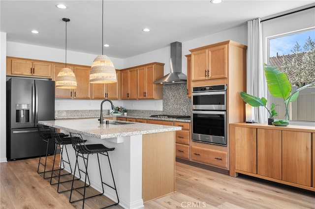 kitchen featuring sink, appliances with stainless steel finishes, wall chimney range hood, a kitchen island with sink, and backsplash