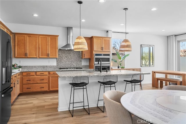 kitchen featuring decorative light fixtures, a kitchen island with sink, wall chimney exhaust hood, black appliances, and light hardwood / wood-style flooring