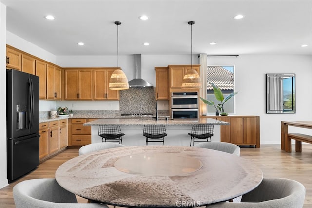 kitchen featuring pendant lighting, a kitchen island with sink, black appliances, light stone countertops, and wall chimney range hood