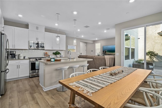 kitchen with light hardwood / wood-style flooring, hanging light fixtures, an island with sink, stainless steel appliances, and white cabinets