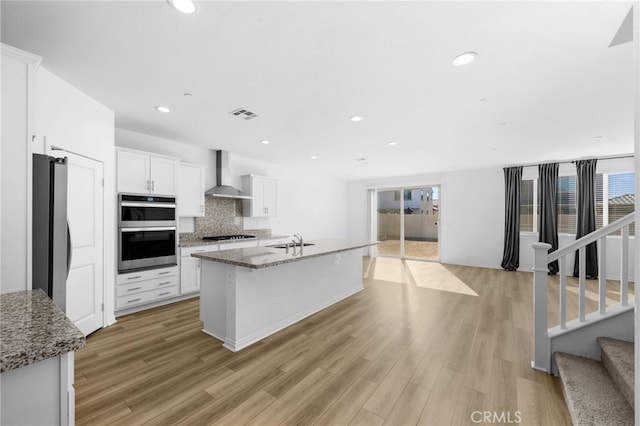 kitchen with white cabinets, appliances with stainless steel finishes, wall chimney range hood, and an island with sink