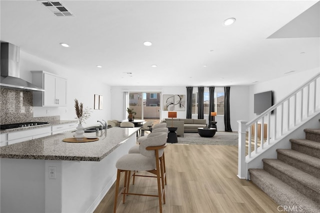 kitchen with light stone countertops, white cabinets, wall chimney exhaust hood, light hardwood / wood-style flooring, and stainless steel gas stovetop