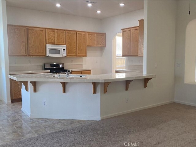 kitchen with light carpet, kitchen peninsula, stainless steel range, tile counters, and a kitchen breakfast bar