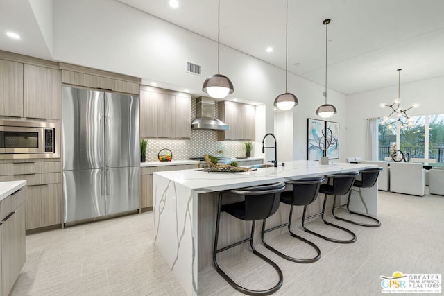 kitchen featuring hanging light fixtures, appliances with stainless steel finishes, a kitchen island with sink, and wall chimney exhaust hood