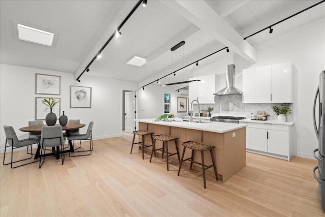 kitchen with pendant lighting, wall chimney range hood, sink, white cabinetry, and a kitchen island with sink