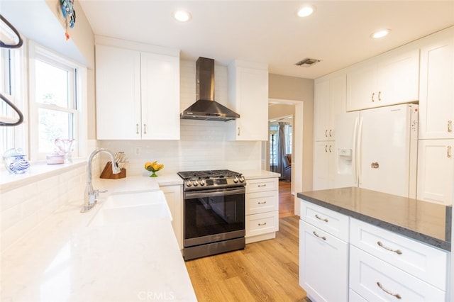 kitchen featuring white fridge with ice dispenser, wall chimney range hood, white cabinets, and stainless steel gas stove