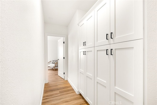 hall featuring a textured wall, light wood-type flooring, and baseboards