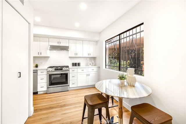 kitchen featuring light wood finished floors, appliances with stainless steel finishes, under cabinet range hood, white cabinetry, and backsplash