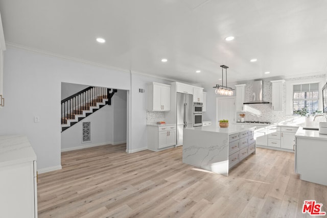 kitchen featuring appliances with stainless steel finishes, wall chimney exhaust hood, a kitchen island, decorative light fixtures, and white cabinetry