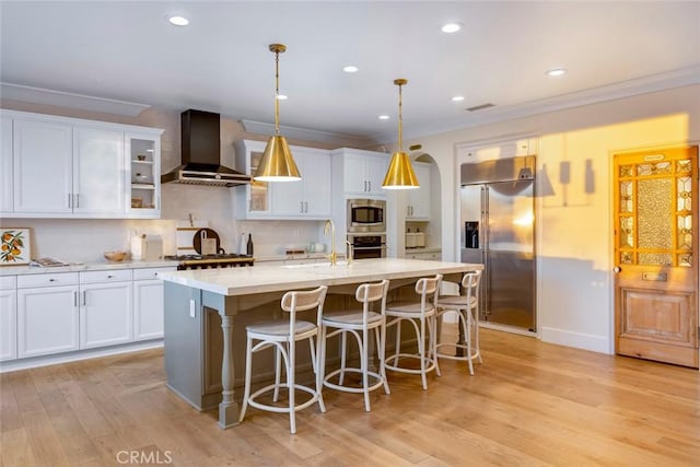 kitchen with wall chimney exhaust hood, an island with sink, white cabinets, and built in appliances