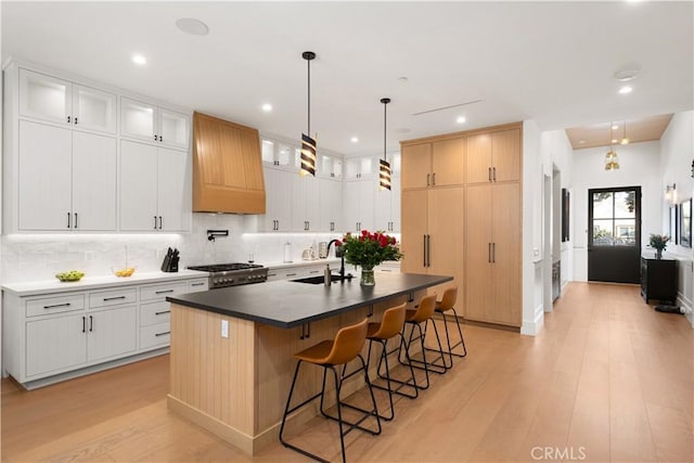 kitchen featuring white cabinetry, an island with sink, custom range hood, stove, and backsplash