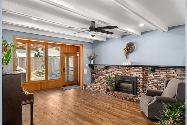 living room featuring ceiling fan, hardwood / wood-style floors, beam ceiling, and a brick fireplace