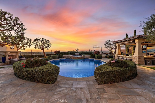 pool at dusk featuring a patio area, an outdoor stone fireplace, and a gazebo