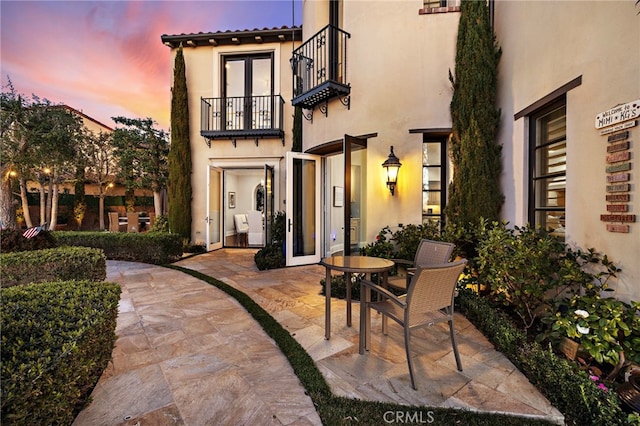 back of house at dusk with a balcony, stucco siding, a patio, and french doors