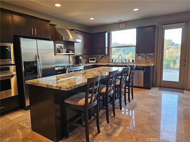 kitchen featuring a breakfast bar, stainless steel appliances, tasteful backsplash, light stone countertops, and wall chimney exhaust hood